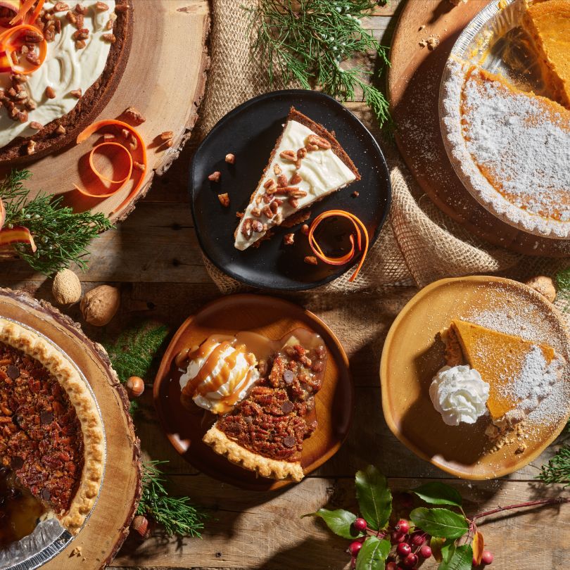 Assorted pies and desserts on a rustic wooden table, featuring a slice on a black plate with nuts and orange peel twists.