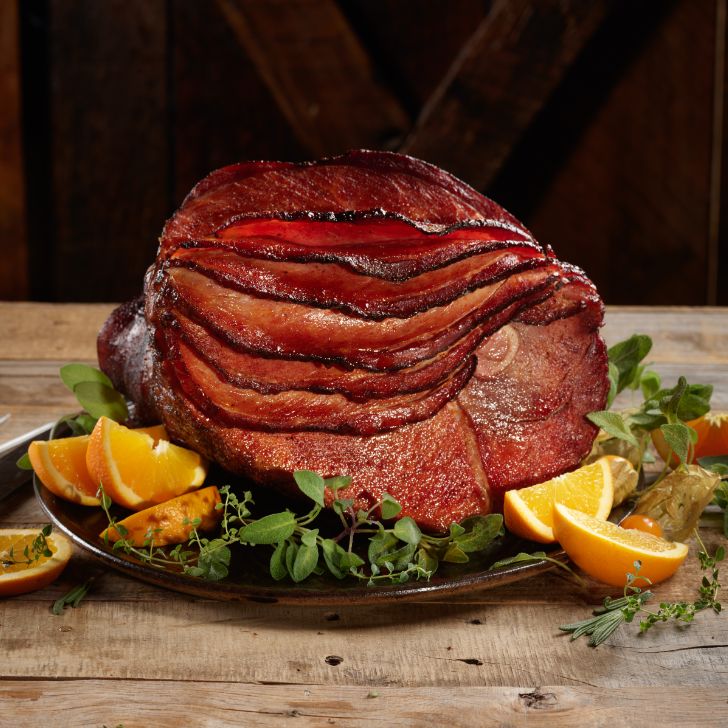 Glazed roast ham piled in slices on a platter with orange wedges and fresh herbs.