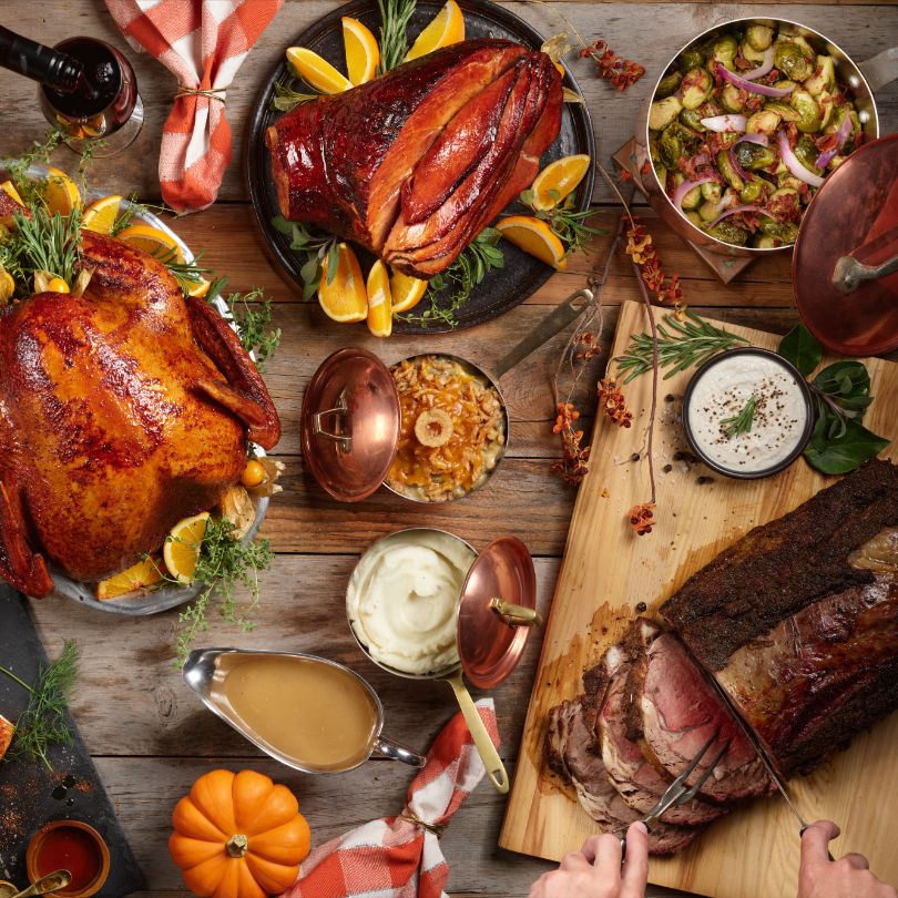 A festive table spread with a whole roasted turkey, sliced beef on a cutting board, and an array of sides and sauces for a holiday meal.