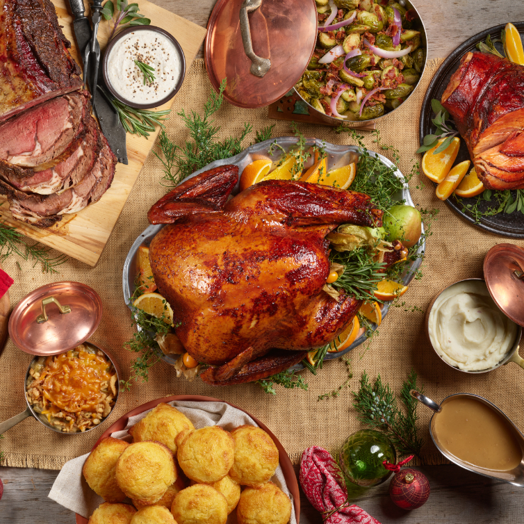 Family-style holiday feast with a glazed roasted turkey centerpiece surrounded by beef, sides, and biscuits on a wooden table.